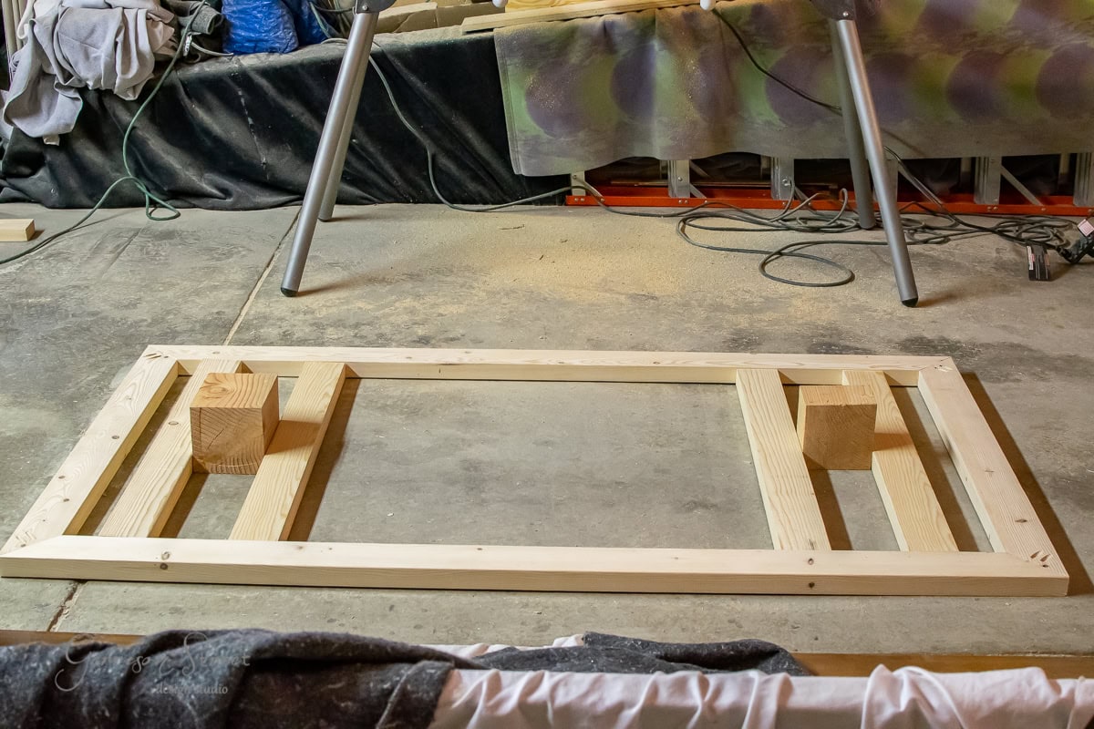 A wooden frame sits on a concrete floor, its structure supported by two large blocks. Tools and materials are scattered in the background, with a partially covered workbench nearby.