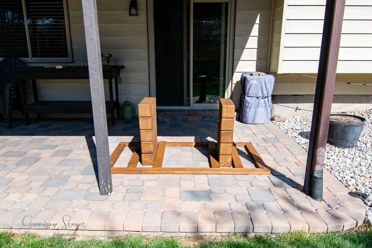 A wooden frame and two stacks of bricks rest on a patio with a stone tile floor. Adjacent to a house with beige siding and a dark door, the scene also features a grill and some gardening tools in the background.