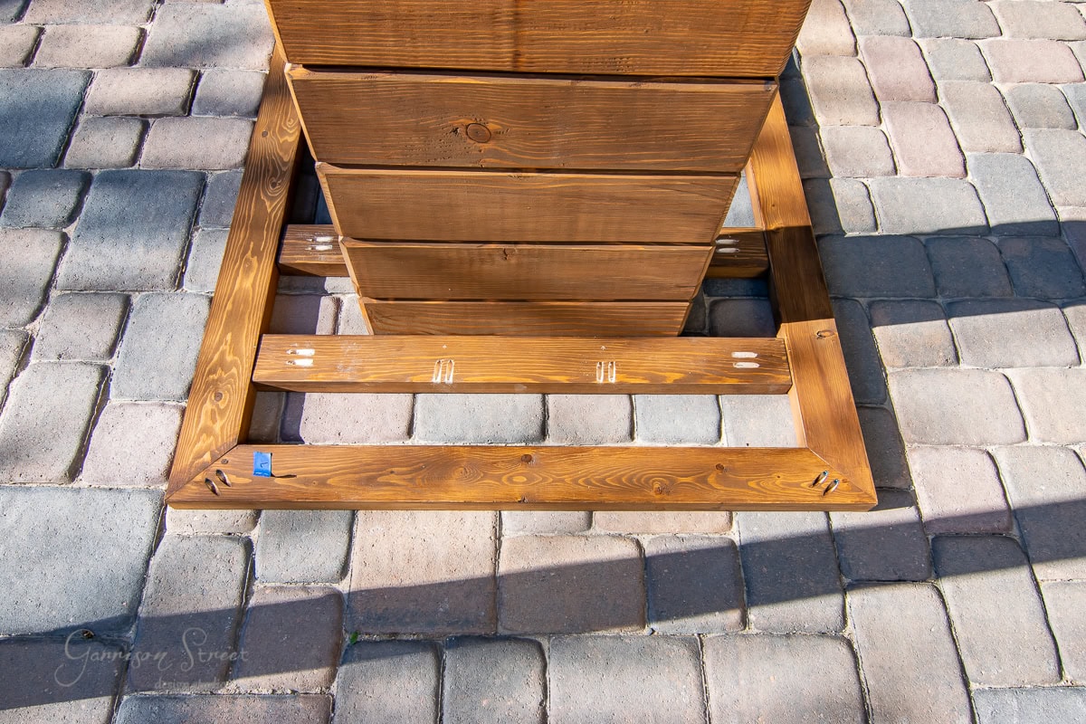 A partially assembled wooden frame rests on a stone-paved surface, viewed from above. The frame showcases metal brackets and screws against the wood's natural finish, with a blue piece of tape marking one corner for alignment.