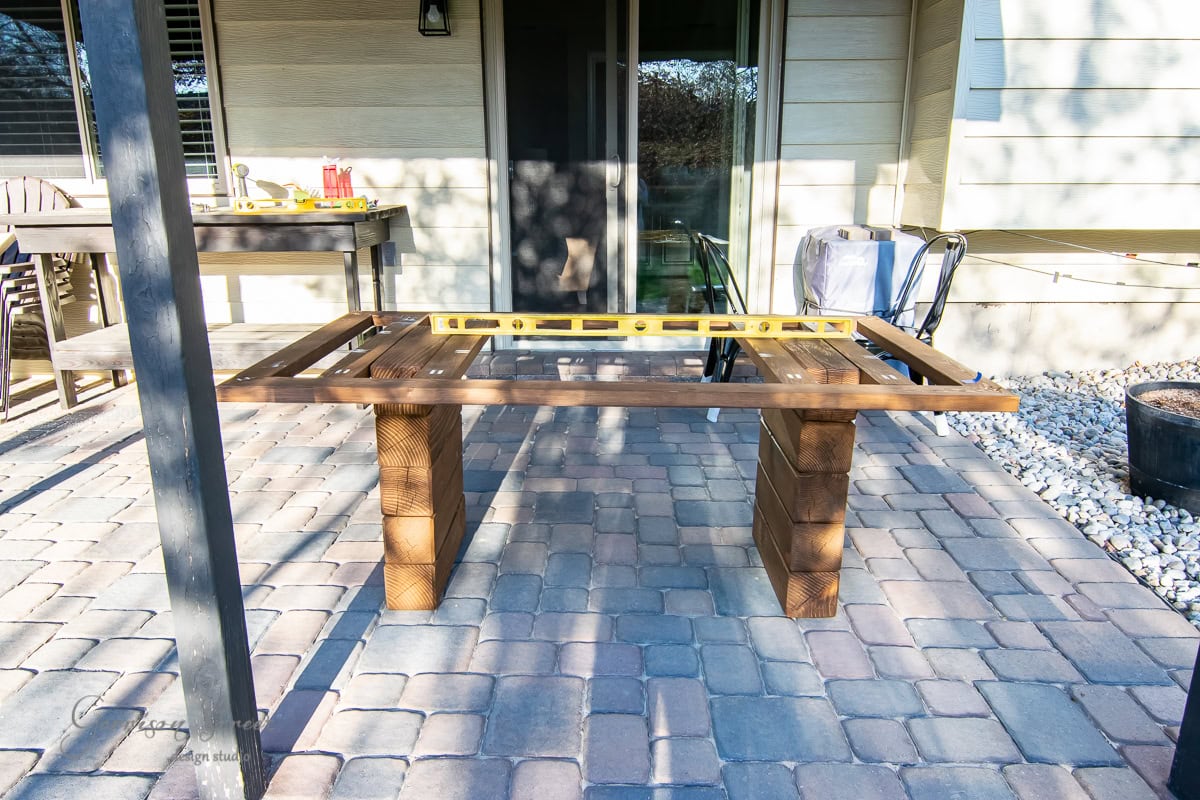 A wooden table frame with rectangular legs sits on the patio, boasting a yellow level on its top. Patio furniture and a grill enhance the setting, while stone flooring and a beige house exterior complete the scene.