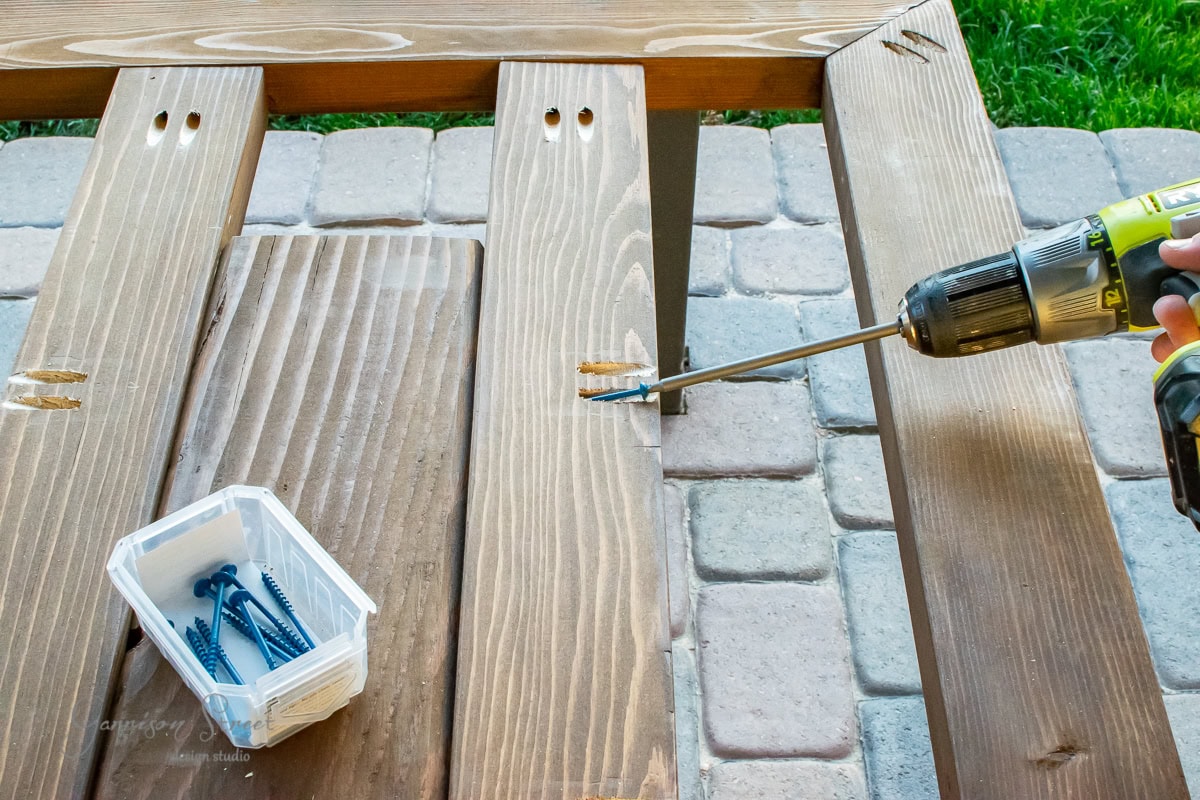 A person is using a drill to assemble wooden planks into a DIY outdoor table on a paved surface. A plastic container holds more screws nearby. The wood has pre-drilled holes, making the project easier. Grass can be seen in the background, adding to the scenic workspace.