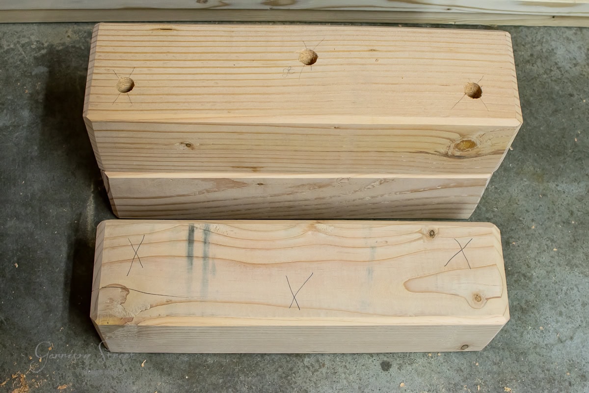 Two wooden beams on a concrete surface. The top beam features three drilled holes, while the lower one displays pencil marks, including several 'X' marks. Both beams showcase natural wood grain patterns.