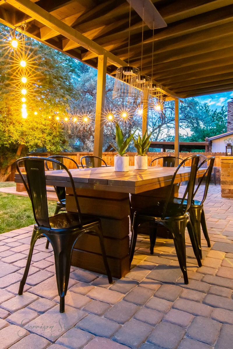 The outdoor patio features a DIY outdoor table set under enchanting string lights. Surrounded by six metal chairs, the table is adorned with potted plants, creating a cozy evening atmosphere against the backdrop of trees in the paved area.