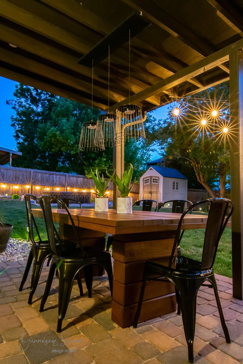 The cozy outdoor patio features a DIY outdoor table set for six, surrounded by metal chairs. Hanging lanterns cast a warm glow over the scene. The backdrop includes trees, a lit fence, and a small shed, all beautifully silhouetted against the twilight sky.