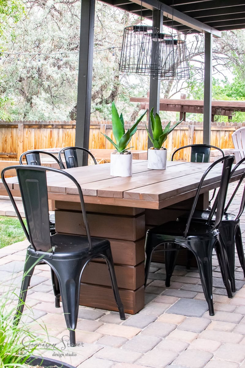 Outdoor patio featuring a DIY outdoor table with black metal chairs on a stone surface. The wooden table hosts two potted plants, while a wire-frame light fixture hangs above. A wooden fence and lush greenery provide a serene backdrop.
