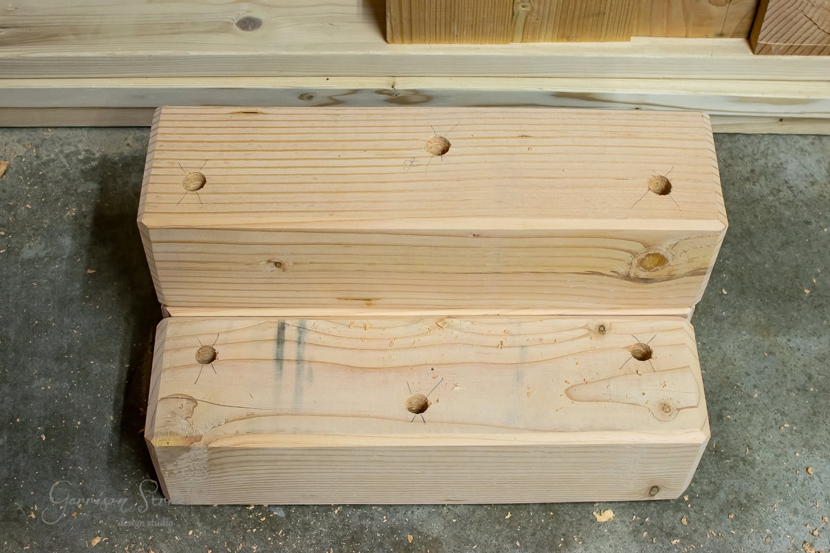 Two rectangular wooden beams with drilled holes rest on a concrete floor. The wood boasts a light natural finish, highlighting its grain patterns and markings. In the background, a stack of similar beams waits to be transformed.