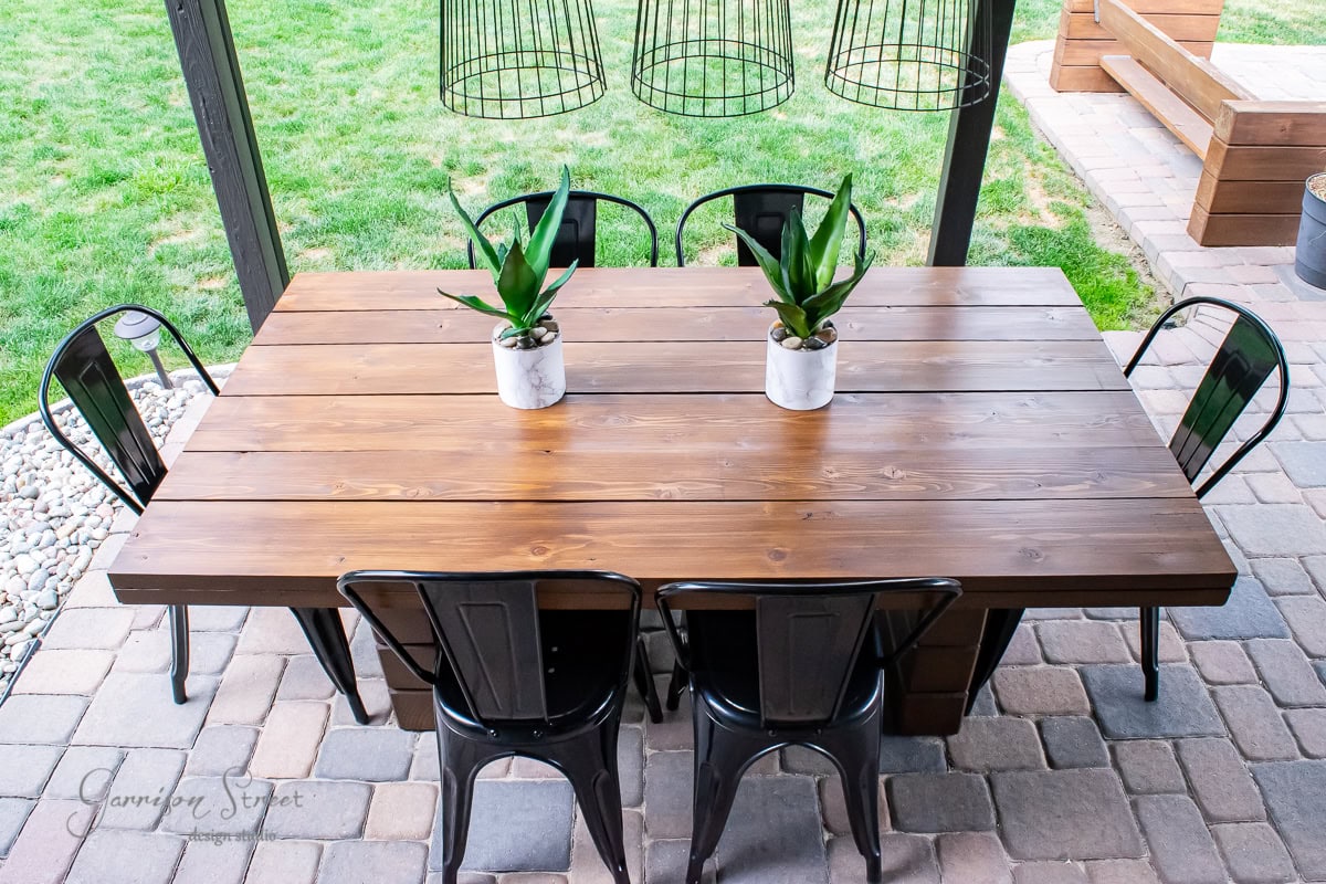 A DIY outdoor dining table crafted from wood is paired with six sleek black metal chairs on a stone patio. Two small potted plants grace the table's center, while wireframe pendant lights hang overhead. In the background, a lush grassy area completes this inviting setup.