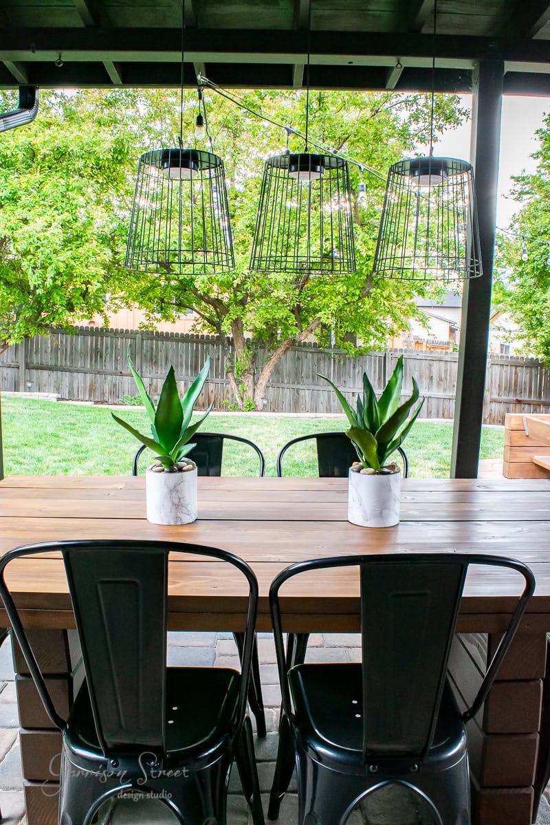 A DIY outdoor patio table is paired with black metal chairs and adorned by two potted plants under three wire-frame pendant lights. The setting is enhanced by a wooden fence and lush green trees in the background, creating a serene and stylish outdoor retreat.