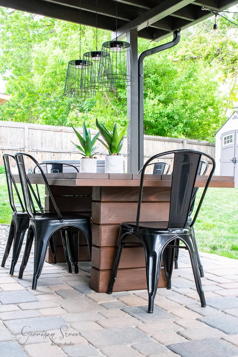 A DIY outdoor table anchors the patio, surrounded by six black metal chairs. On its surface rest two potted plants, while overhead wireframe pendant lights cast a soft glow. In the background, a grassy yard beckons, bordered by a sturdy wooden fence.