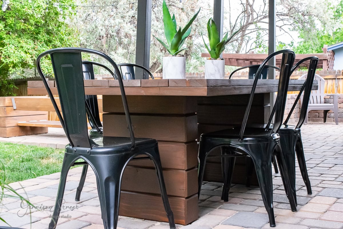 A DIY outdoor table crafted from wood features two small potted plants atop its surface, set on a stone patio. Four black metal chairs encircle the table, while lush green plants and trees provide a serene backdrop.