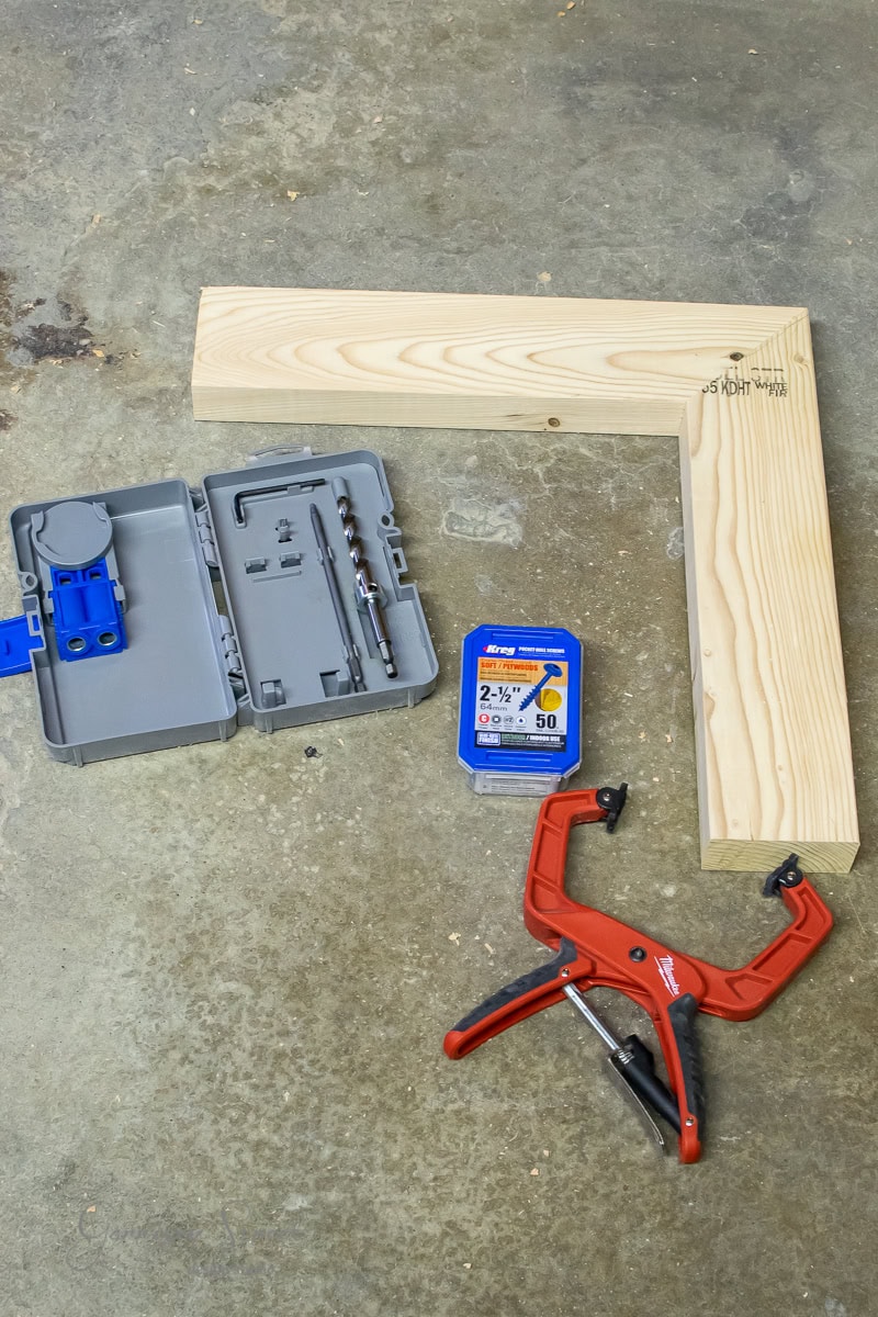 Carpentry tools and supplies for building your own DIY outdoor table are neatly arranged on a concrete floor, featuring a wooden right angle, a grey drill bit kit, a red clamp, and a box of 2-1/2 inch screws.