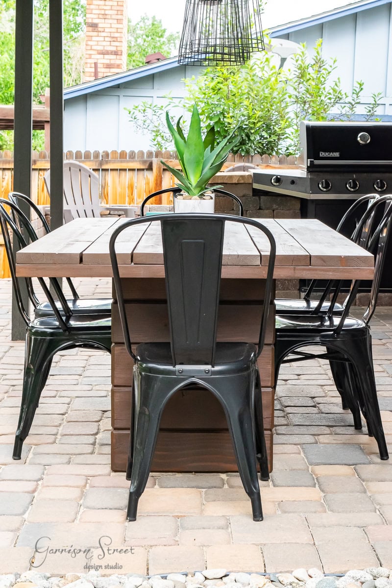 The outdoor patio features a DIY wooden dining table paired with black metal chairs on a stone floor. A small potted plant is centered on the table, adding charm to the setup. In the background, a barbecue grill complements the scene, enveloped by lush greenery.