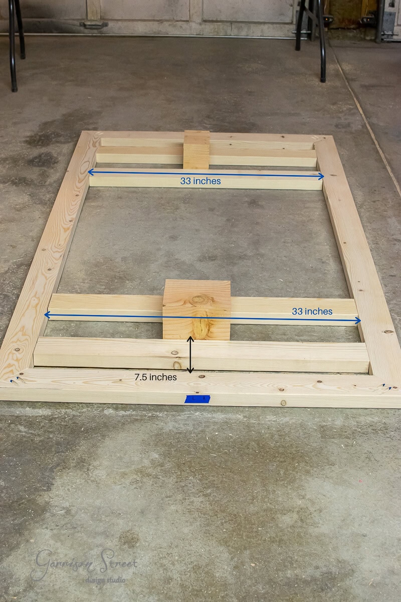Wooden frame on a floor made of natural, unpainted lumber. Two horizontal support beams align inside, with blue tape marking measurements of 33 inches and 7.5 inches. A block sits at the center intersection, and tools are scattered in the background.
