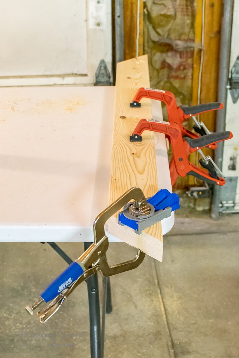 A piece of wood is clamped to a workbench using several red and blue clamps. The setup resides in a workshop filled with tools and materials.