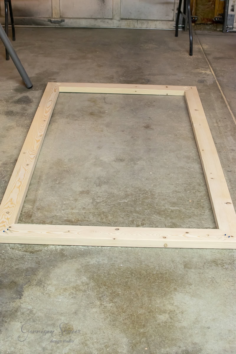 A wooden rectangular frame lies on the concrete floor in a workshop. Several tools and a chair are visible in the background.