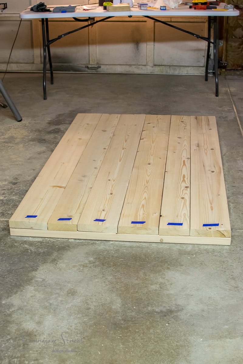 A partially assembled wooden pallet lies on a concrete floor in a workshop. A table with various tools and materials is visible in the background. The pallet consists of several wooden planks placed parallel to each other.