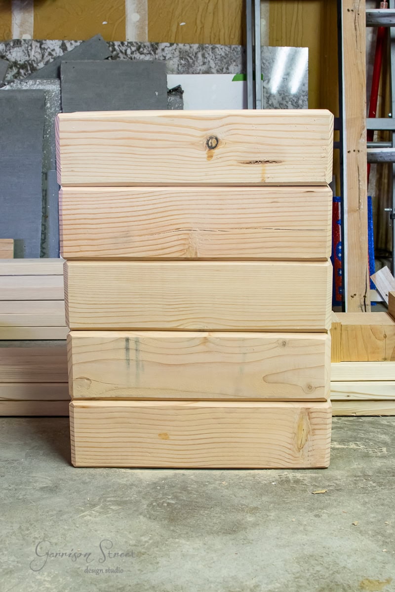 A stack of six wooden planks is neatly arranged vertically against a workshop wall, surrounded by various tools and materials. The light-colored planks display their natural wood grain beautifully.