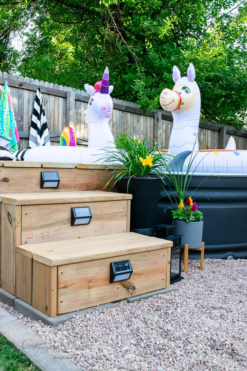 In the backyard, a stock tank pool is adorned with inflatable unicorn floats. Wooden steps lead up to the pool, surrounded by colorful towels, potted plants, and decorative flowers. A fence and lush greenery provide a charming backdrop to this idyllic retreat.