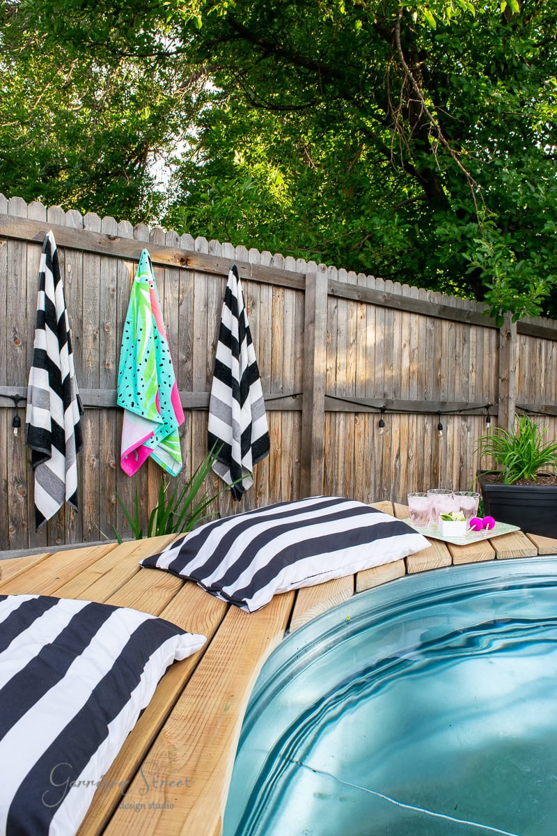A cozy backyard scene featuring a charming stock tank pool with a wooden deck. Striped black and white pillows and towels are placed around the pool. A fence and green trees provide privacy. Drinks and a flower rest on the deck, enhancing the relaxing atmosphere.