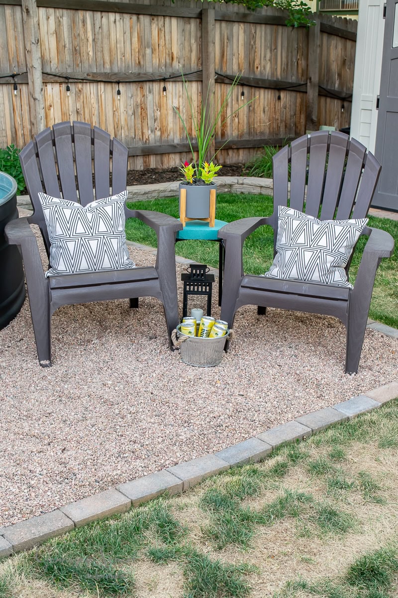 Two gray Adirondack chairs with patterned cushions sit on a gravel area. Between them is a turquoise table holding a potted plant. A small lantern and bucket with drinks rest on the ground, next to a stock tank pool, all set against a wooden fence backdrop.