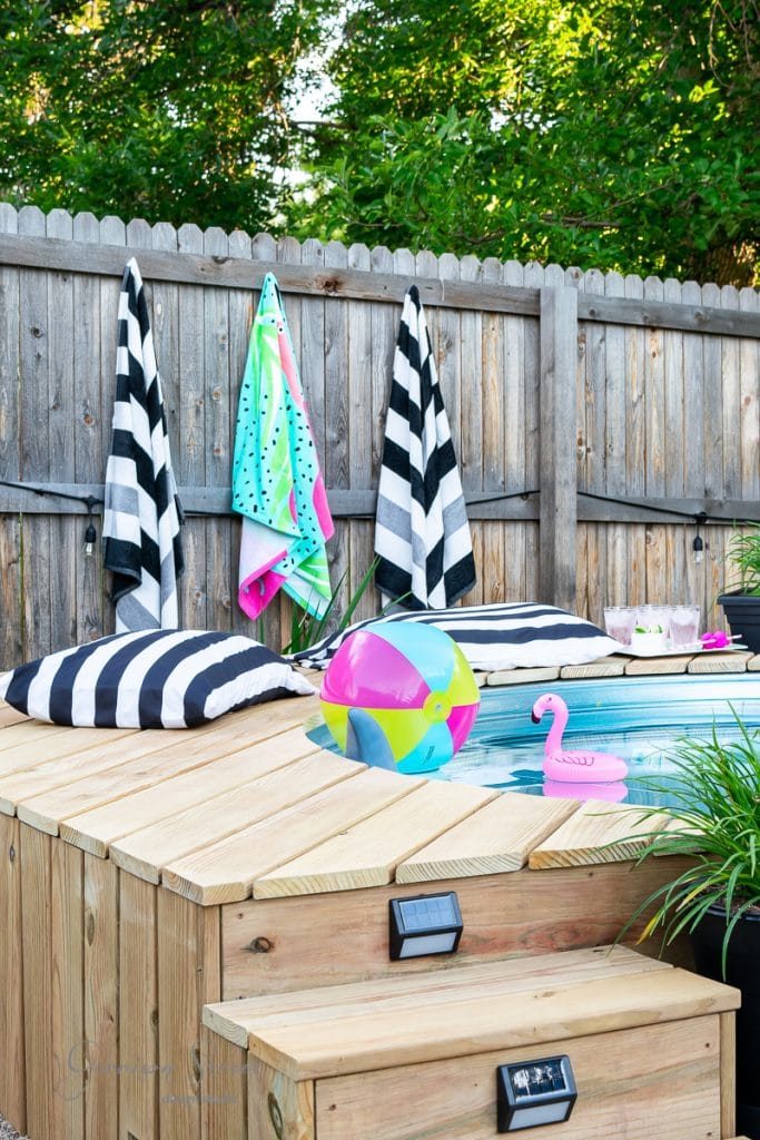 Home A backyard pool with a wooden deck, featuring black and white striped towels hanging on a fence. A colorful beach ball and pink flamingo float are in the pool, with black and white striped pillows nearby. Lush green trees are in the background.