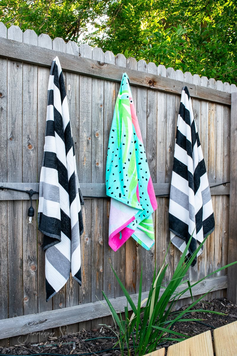 Three beach towels hang on a wooden fence near a vibrant stock tank pool in a garden. The left and right towels feature black and white stripes, while the middle towel showcases a colorful watermelon design. Lush greenery surrounds the serene oasis.