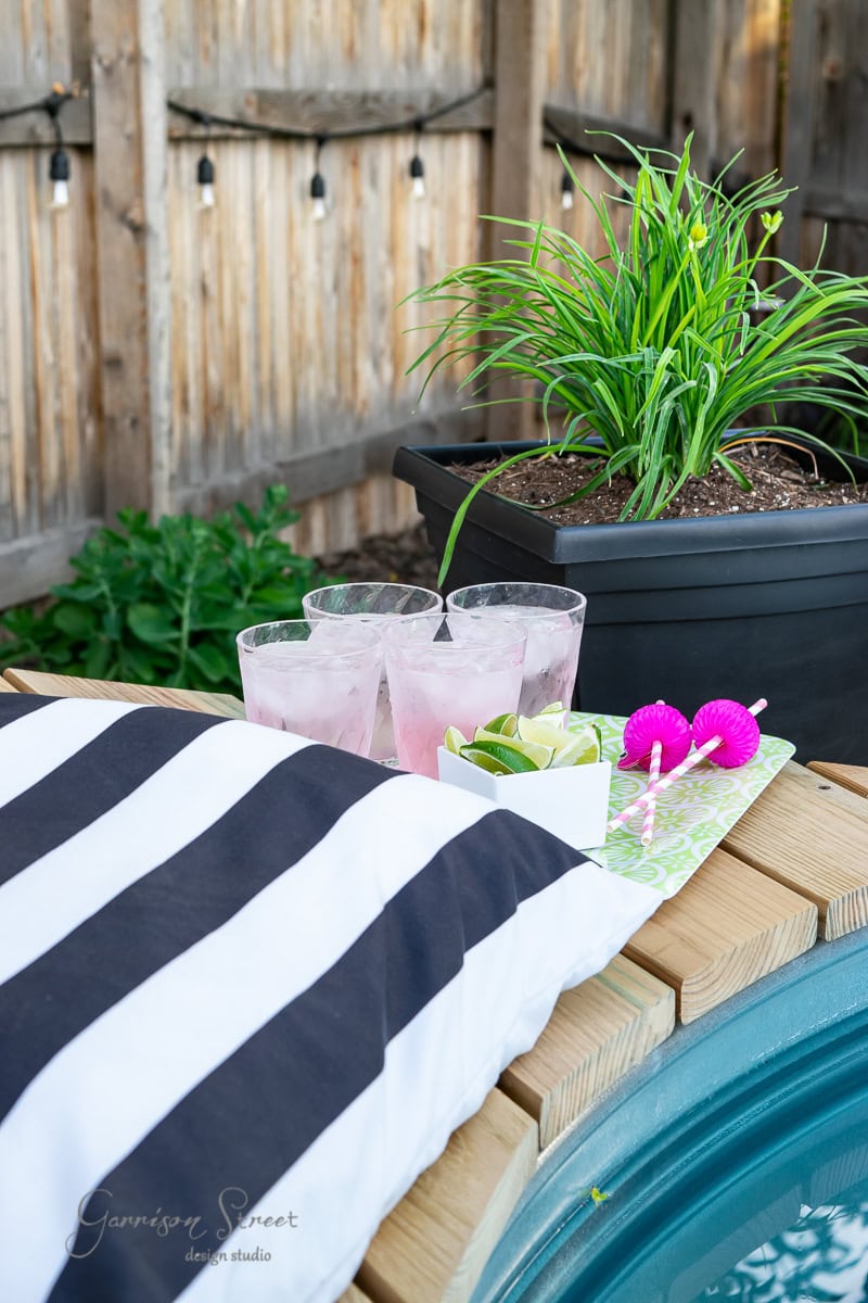 A cozy outdoor setting features a striped black and white pillow by the stock tank pool. A tray holds three pink drinks with ice and a small dish of lime wedges. A potted plant sits in the background, next to a wooden fence adorned with string lights.
