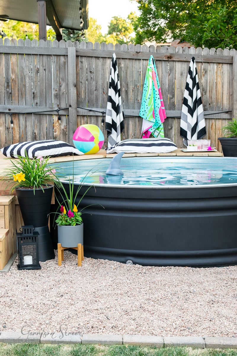 A backyard scene features a round stock tank pool surrounded by wooden decking and pebbles. Towels and cushions with black and white stripes hang on the fence. A colorful beach ball floats in the water, while potted plants and a lantern add charm to the area.