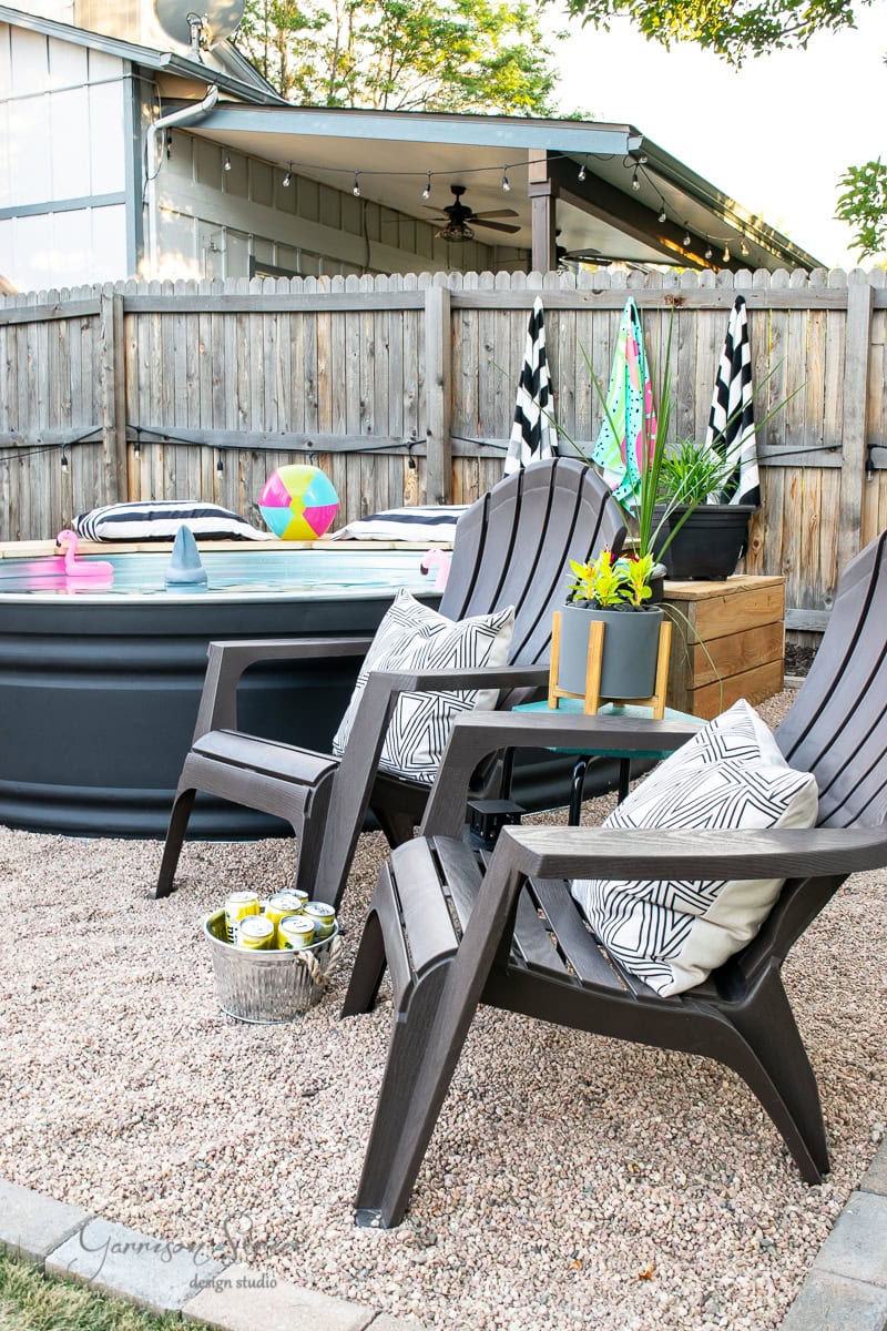 Patio with two dark Adirondack chairs and patterned cushions, facing a stylish stock tank pool. A small table holds a potted plant, with a bucket of canned drinks on the ground. Colorful pool toys are visible, and a wooden fence encloses the area.