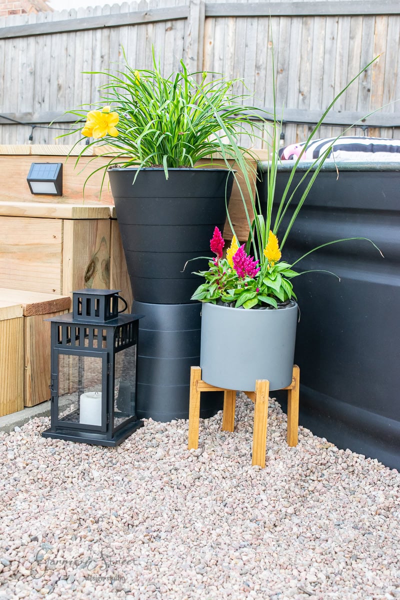 A small garden space features a black lantern on gravel, a tall black planter with yellow flowers, and a gray planter on a wooden stand filled with colorful blossoms. A charming stock tank pool adds to the ambiance, while wooden seating and a fence complete the picturesque backdrop.