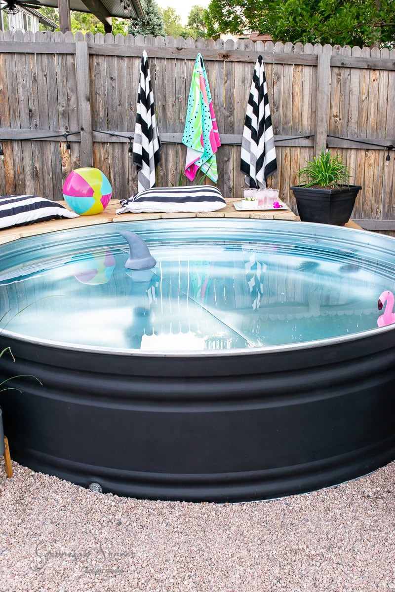 A round, stock tank pool with clear blue water stands on a gravel surface. Three striped towels hang on a wooden fence in the background. A colorful beach ball and a small pink toy float gently in the water. Two striped cushions rest invitingly on a nearby bench.