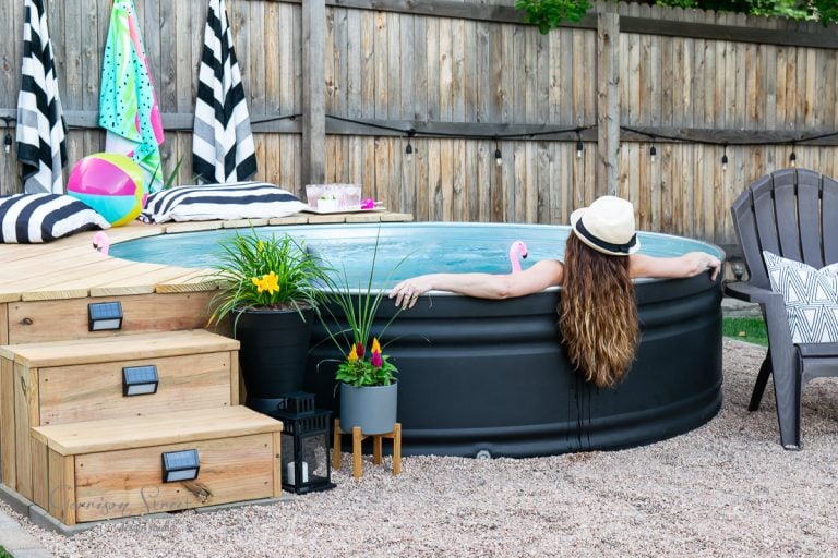 A person in a hat relaxes in a backyard pool, resting their arms on the edge. The scene includes colorful towels, potted plants, and pool toys. Wooden steps lead to the pool, and a wooden fence surrounds the yard.