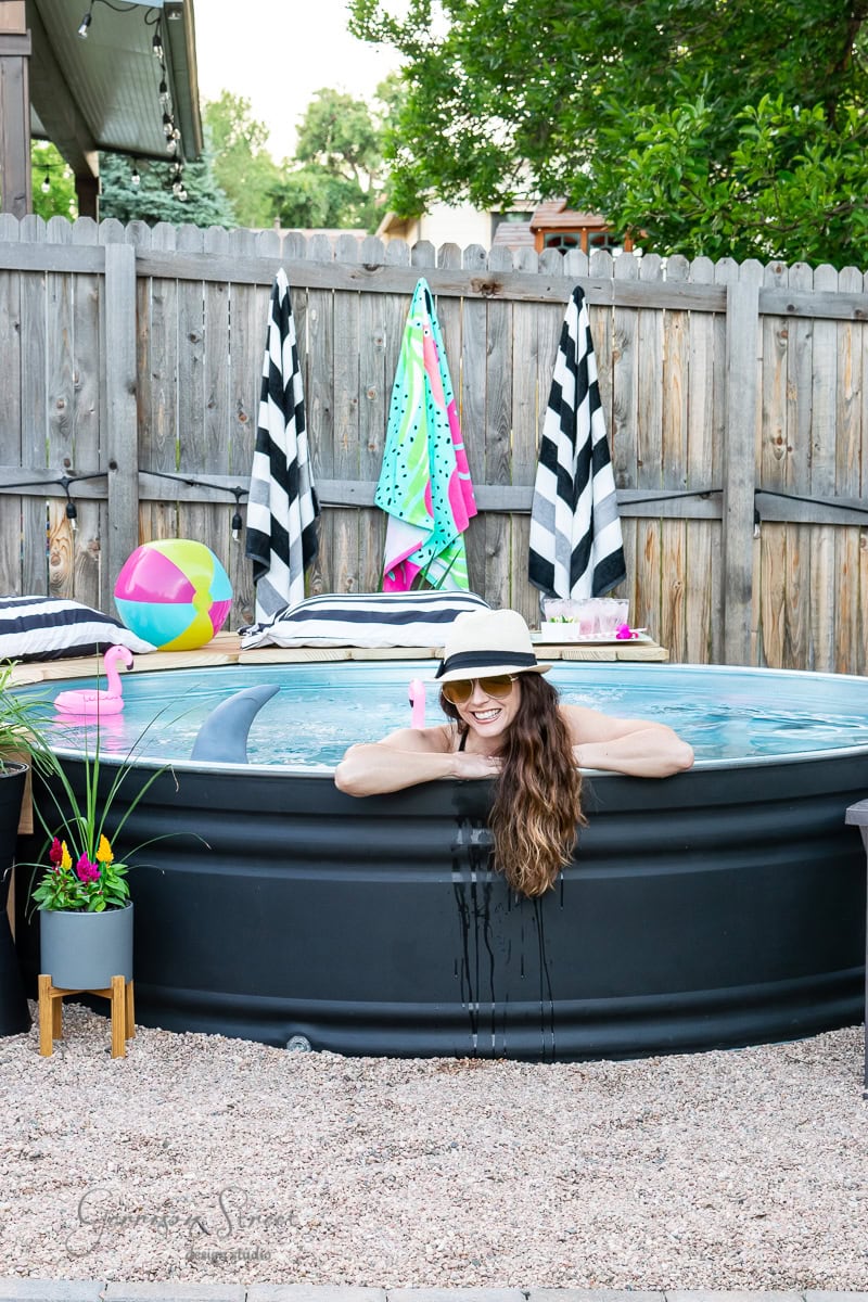 A woman in a sunhat leans on the edge of a cozy stock tank pool, smiling. The area is surrounded by gravel, with colorful towels and a beach ball in the background. Potted flowers and inflatable flamingos add charm, while a wooden fence encloses this picturesque scene.