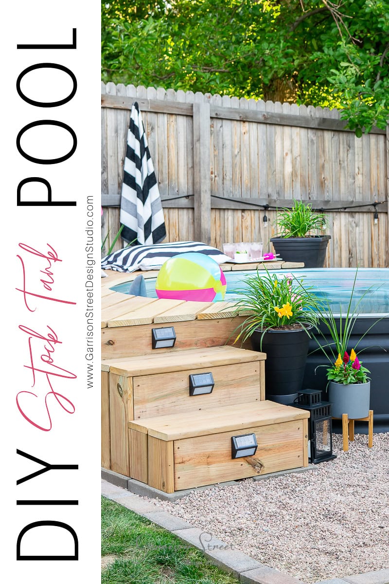 In a charming backyard scene, a stock tank pool is nestled within a wooden deck adorned with various potted plants. Nearby, a colorful beach ball and black-and-white striped towel add a playful touch, while a wooden fence and lush greenery provide the perfect backdrop for this DIY oasis.