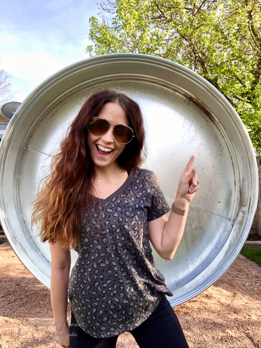 A woman with long hair, wearing sunglasses and a patterned shirt, smiles and points upwards. She stands in front of a large, round metal stock tank pool, with green trees in the background.