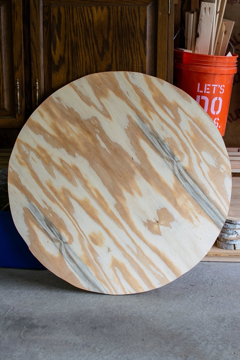 A large, round piece of plywood leans against a wooden cabinet. A red bucket and various pieces of wood lie in the background.