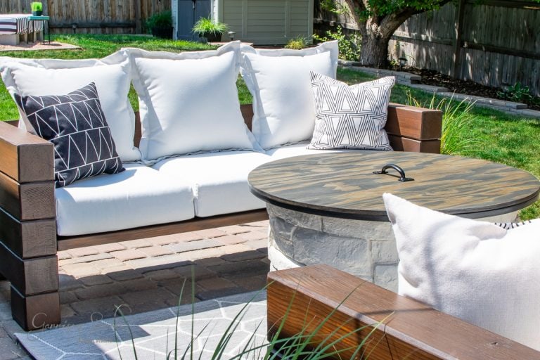 A cozy outdoor patio features a white cushioned wooden sofa with decorative black and white geometric pillows. In front, a round wooden coffee table with a stone base sits on a brick surface. The background shows a green lawn and wooden fence.