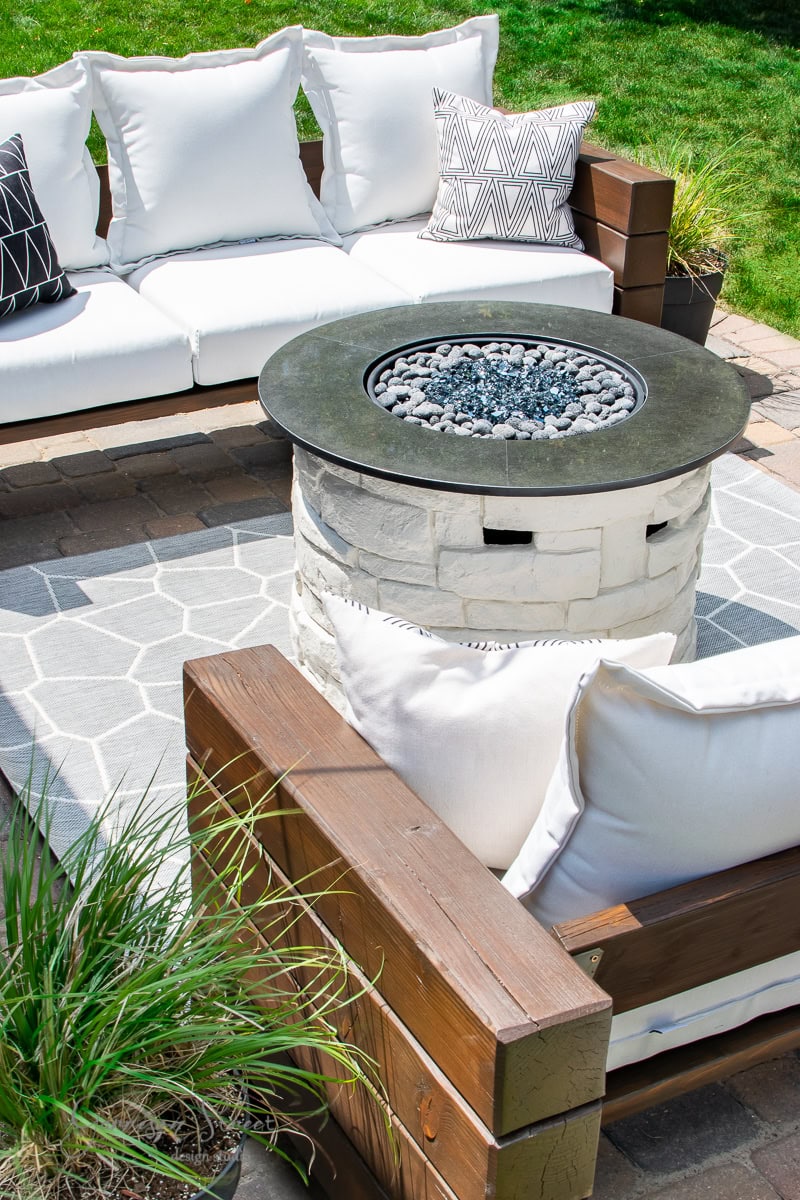 A cozy outdoor seating area with white cushioned sofas and geometric-patterned cushions surrounds a round propane fire pit on a tiled patio. Green grass and ornamental grasses add a touch of nature to the scene.