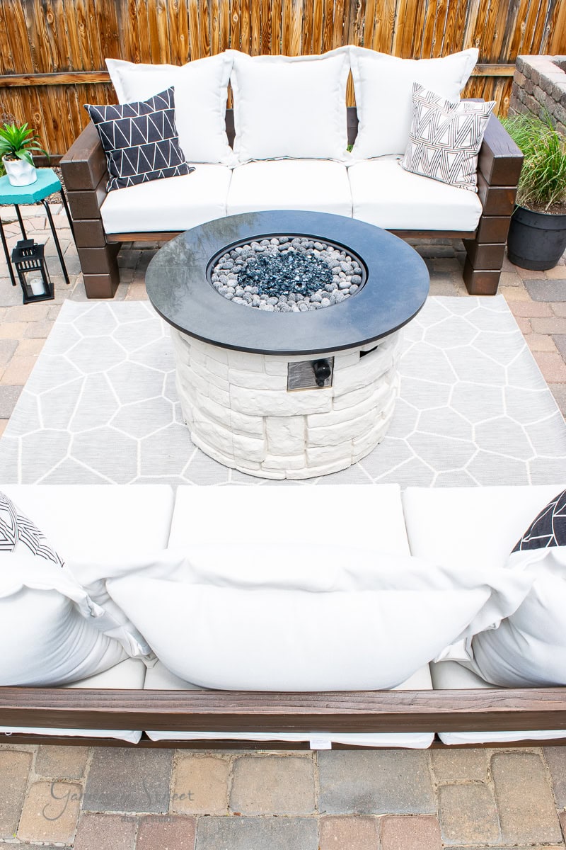 Outdoor patio featuring a propane fire pit table surrounded by two white cushioned sofas. The area boasts geometric patterned cushions and a gray rug on stone pavers. Small plants and a lantern adorn the side table.