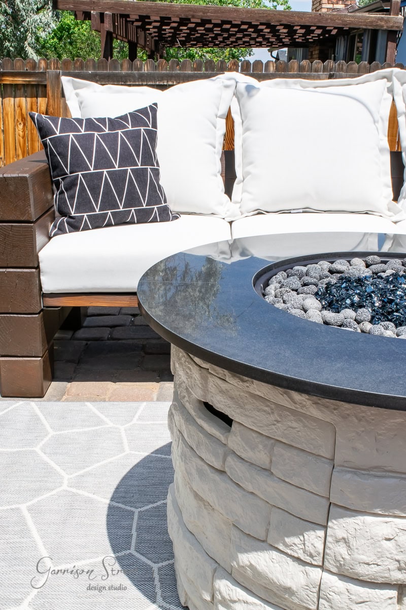 The outdoor patio features a stone fire pit and round glass table. White cushioned seating boasts a black geometric-patterned pillow. A wooden fence and pergola offer a cozy atmosphere, with an optional wood cover for the fire pit adding versatility.