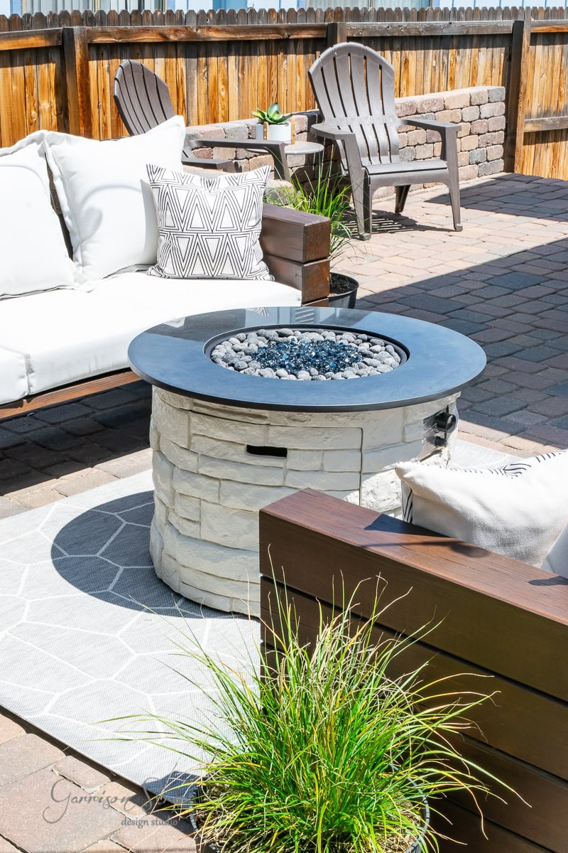 A cozy outdoor patio features a round propane fire pit with black pebbles, surrounded by white cushioned seating and wooden chairs. A gray patterned rug and potted plants add to the inviting atmosphere, while a DIY wooden cover cleverly conceals the fire pit when not in use.