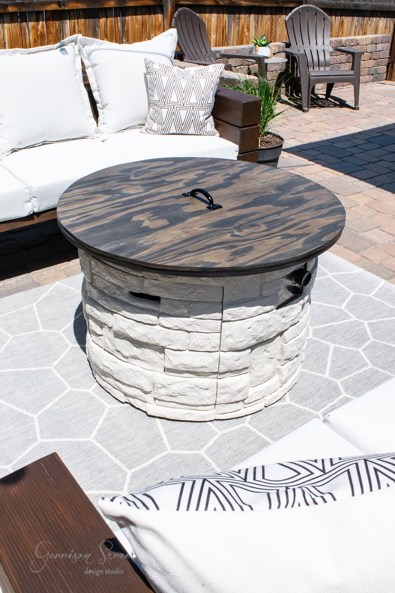 A round outdoor propane fire pit with a wooden cover sits on a patterned rug. Surrounding it are white cushioned seating and a few wooden chairs. The scene is set on a stone patio with a wooden fence in the background.