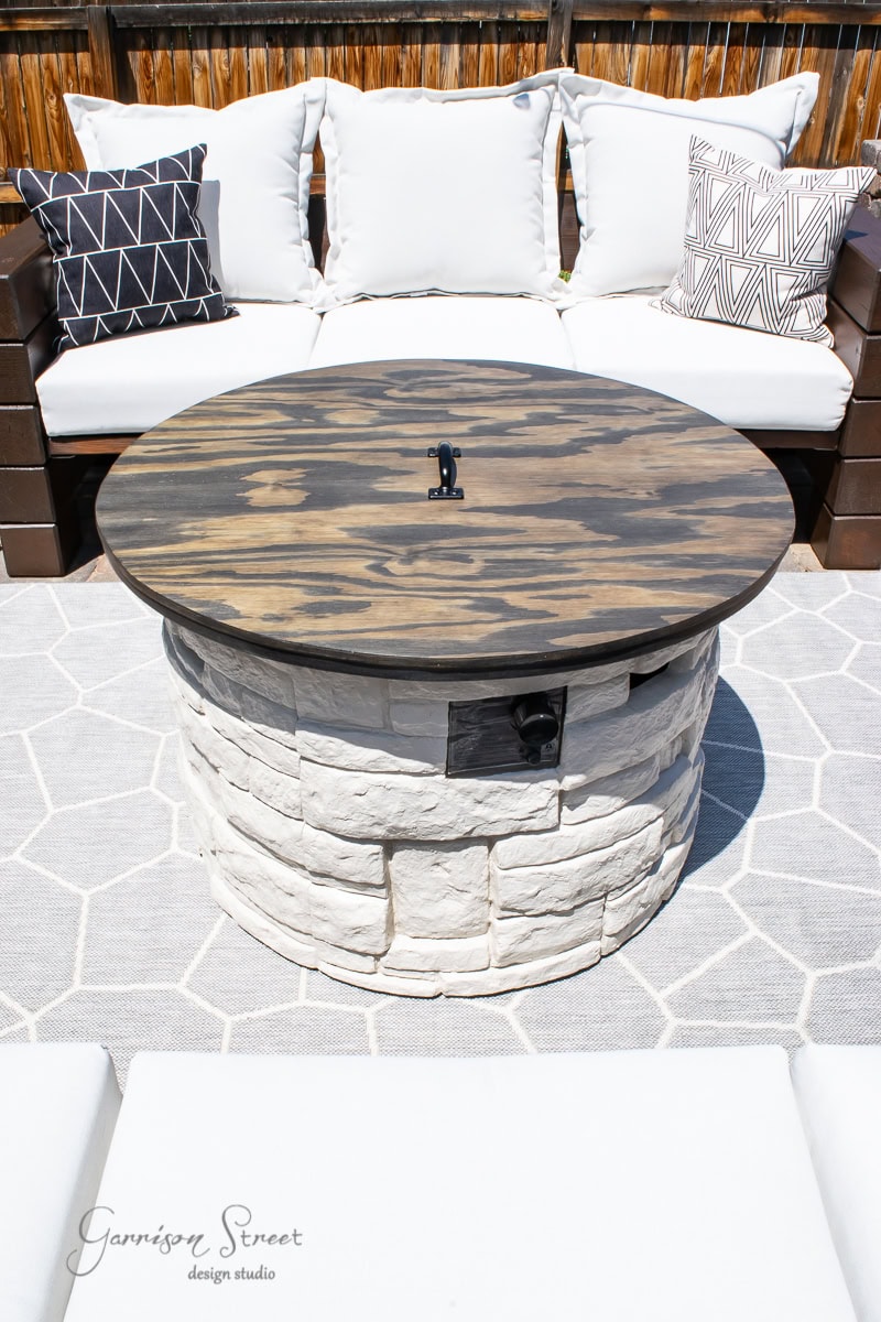 A stone fire pit with a custom DIY wooden cover sits gracefully on a hexagonal-patterned outdoor rug. It's surrounded by a cushioned sectional sofa adorned with black and white geometric patterned pillows, all set on a cozy wooden deck.