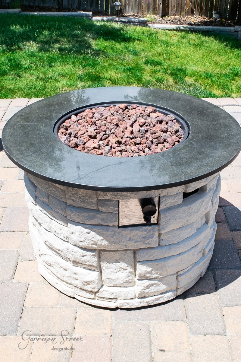 Stone fire pit with a round, dark stone top filled with red lava rocks. It stands on a patio with gray paving stones, surrounded by green grass. A control panel is visible on the side.
