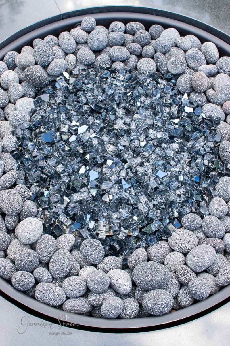 A circular propane fire pit with gray pebbles encircling the edge and a center filled with reflective glass pieces offers a stunning glow for any backyard gathering.
