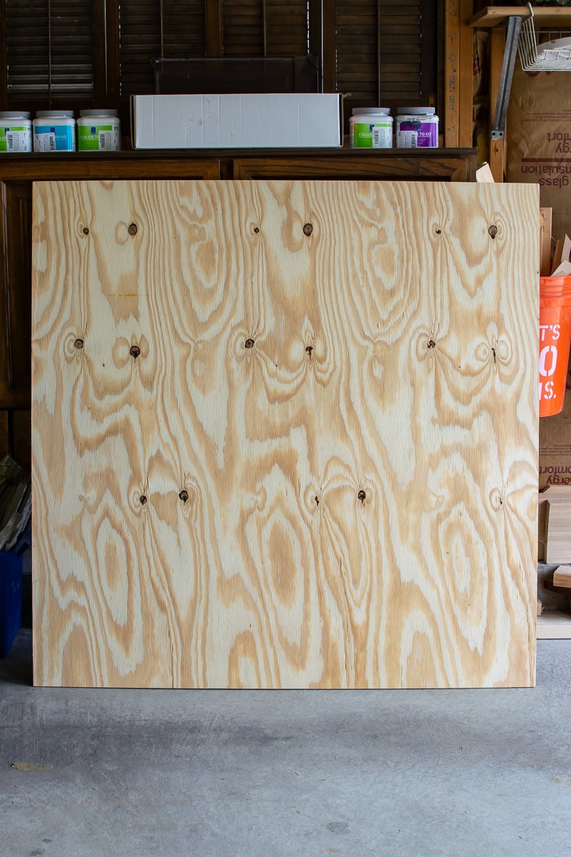 A large sheet of plywood stands upright in a dimly lit garage. The wood showcases a wavy grain pattern with dark knotholes. Shelves in the background hold various colorful cans, hinting at the creative projects underway.