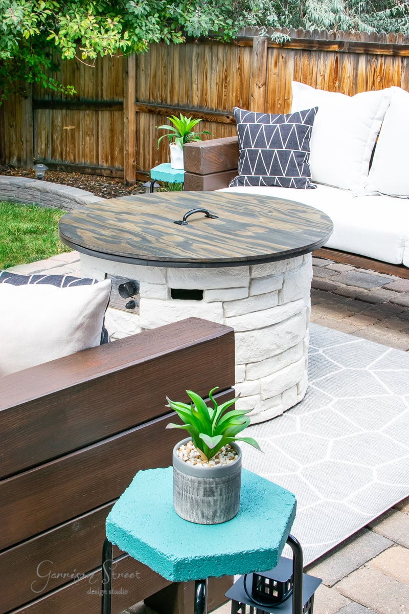 This cozy outdoor patio features a DIY white brick fire pit with a circular wooden cover. Cushioned seating surrounds it, adorned with geometric-patterned throw pillows. A small blue table holds a potted plant, and the area is bordered by a wooden fence and lush greenery.