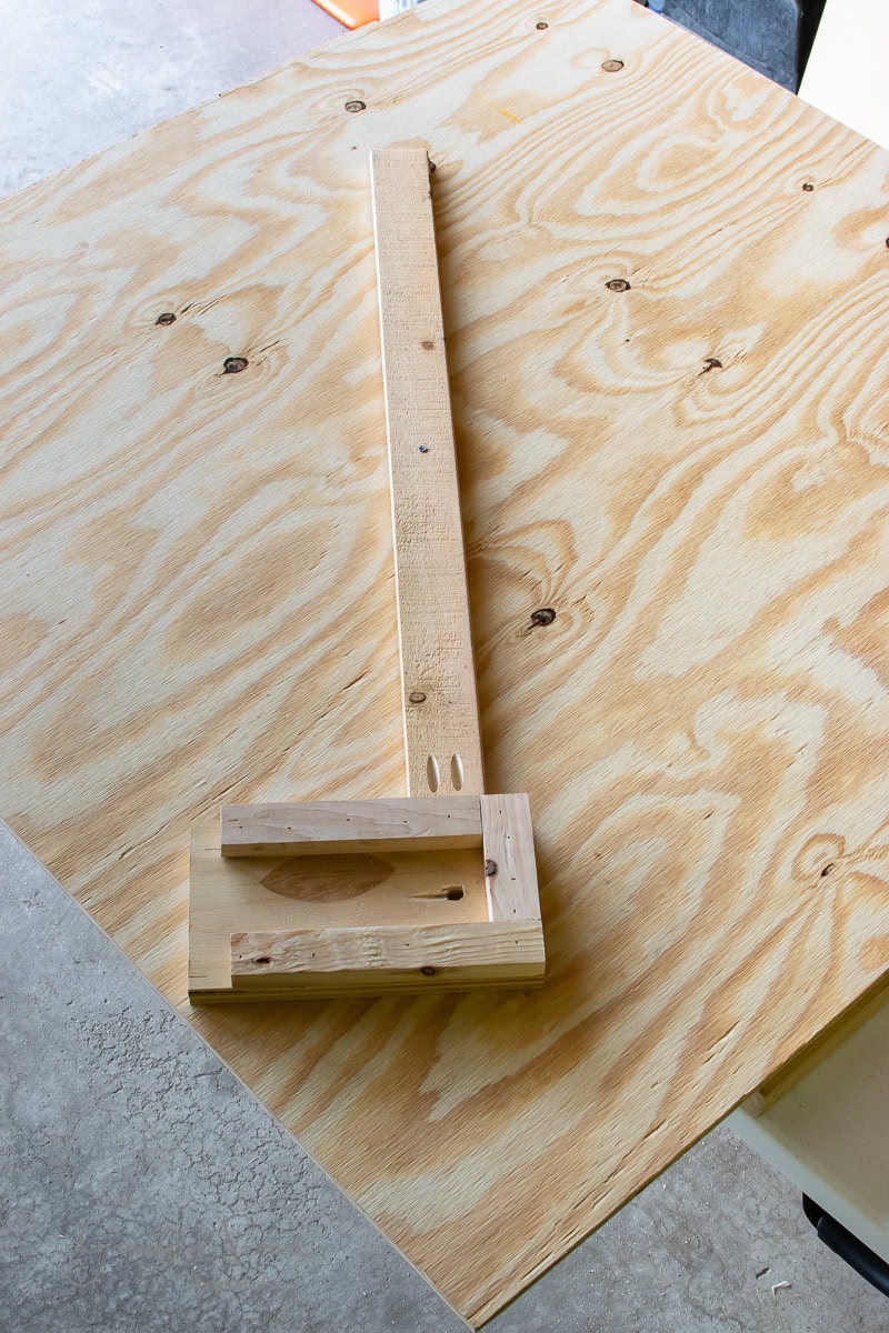 A homemade wooden track saw guide rests on a large sheet of plywood. The guide, crafted from two pieces of wood joined at a right angle, ensures straight cuts. The plywood's grain pattern complements the workshop floor.