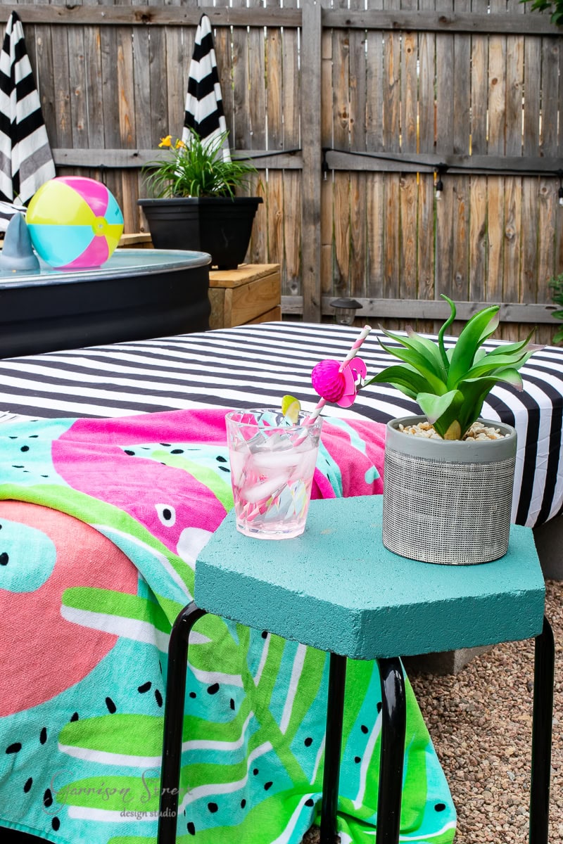 A colorful outdoor scene features a striped sunbed with a watermelon-themed towel draped over it. A small teal DIY table holds a potted plant and a glass with a pink flamingo stirrer. In the background, you can see a pool with a beach ball floating on its surface.