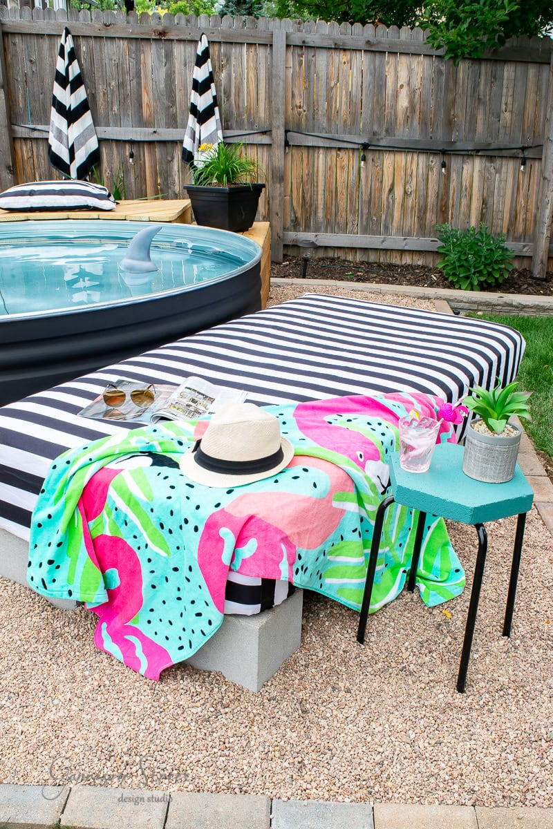 A backyard pool area features a black inflatable pool beside a chic sunbed adorned with a colorful flamingo towel. A straw hat, sunglasses, and magazine rest on the bed, while nearby a small table holds potted plants. Black and white striped umbrellas provide shade.